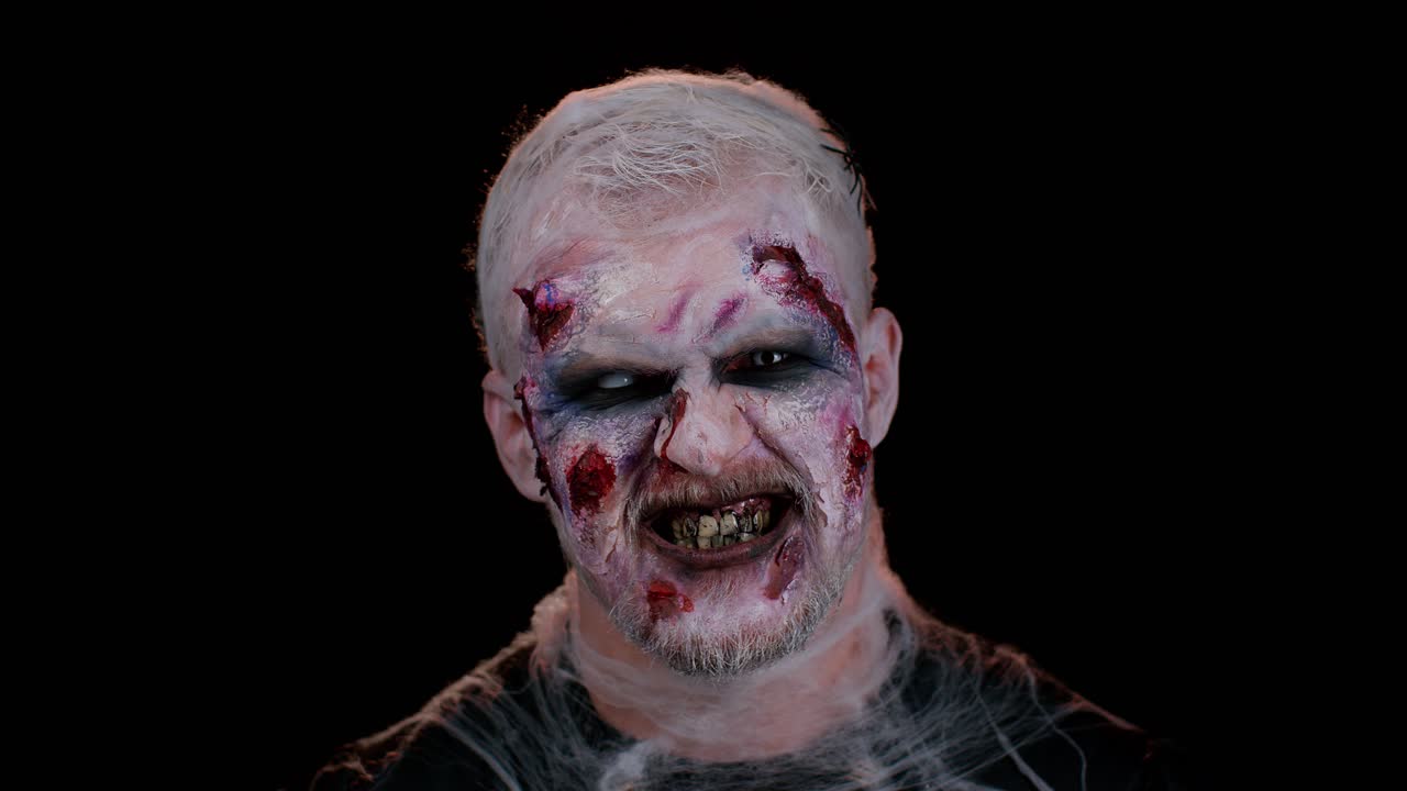 Zombie man with wounds scars and contact lenses looking at camera clicks his teeth trying to scare