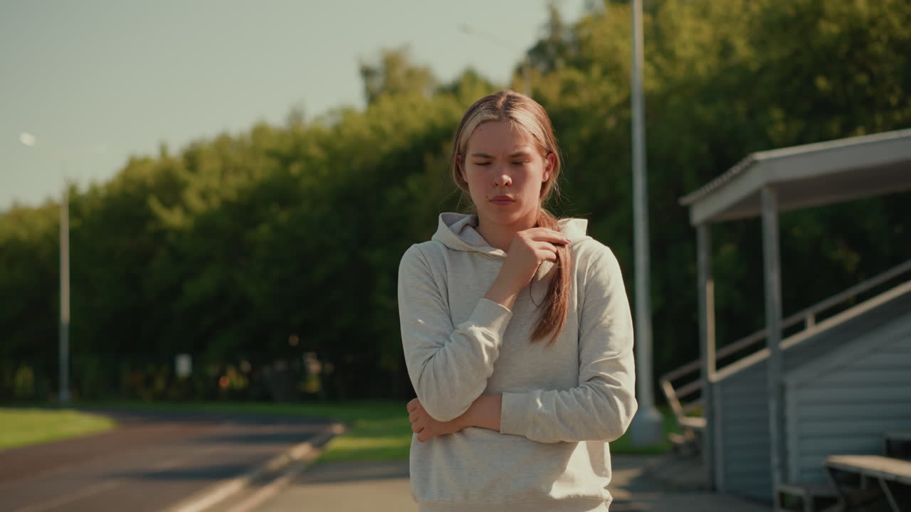 Young woman in casual hoodie standing thoughtfully in empty stadium, gently touching her hair while sunlight casts a warm glow on her face and background trees