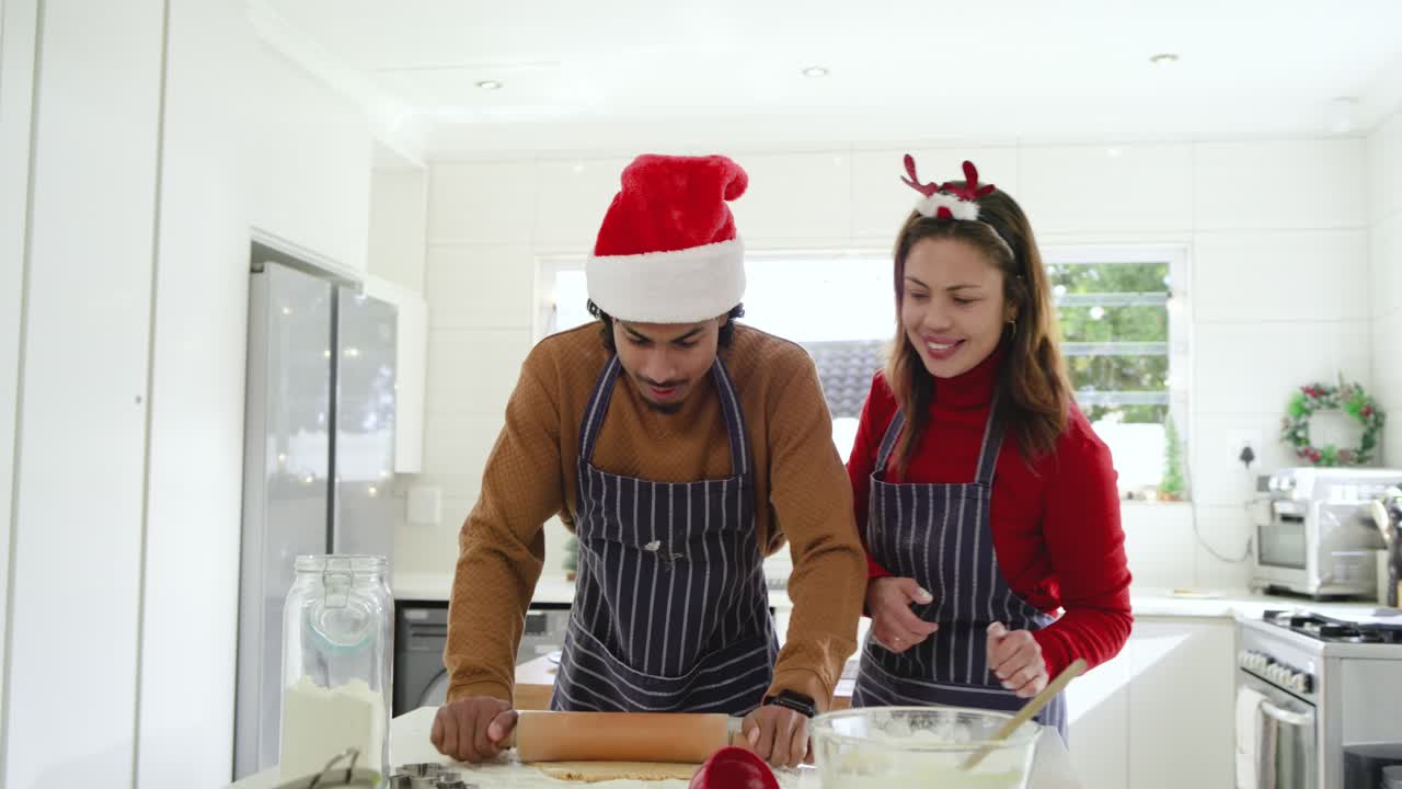 Diverse couple draping arm around shoulder and preparing cookies with rolling pin at kitchen island