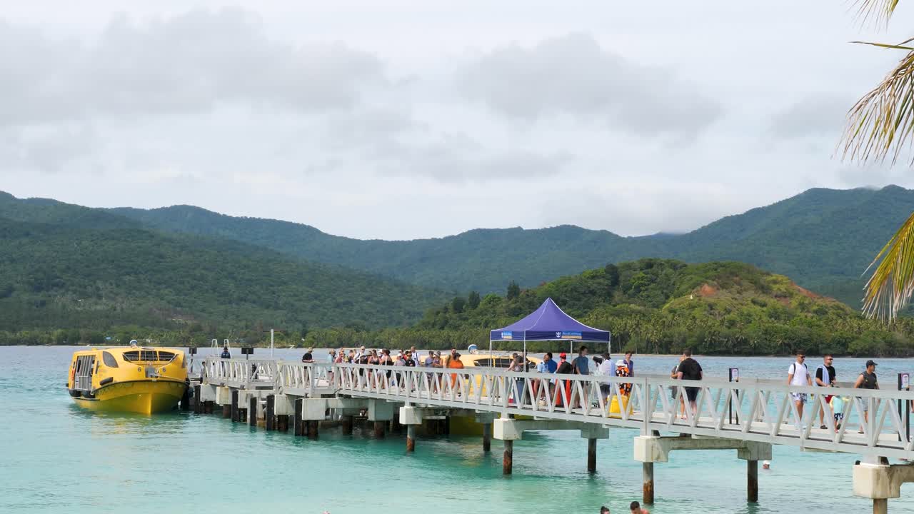 Crowds of people walking on a pier towards a yellow boat at a tropical island