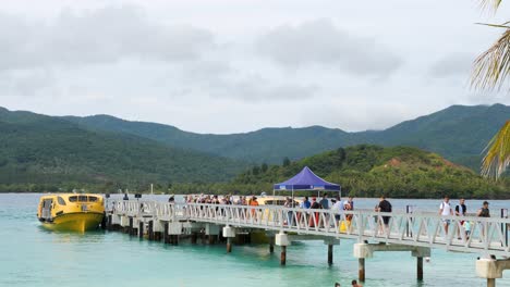crowds of people walking on a pier towards a yellow boat at a tropical island