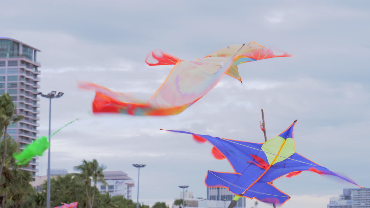 Colorful kites are displayed on bamboo poles at the foreground, and in the background beachgoers are swimming, sailing, and strolling at the beach in Pattaya at Chonburi province in Thailand