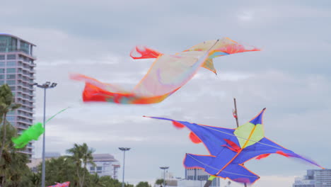 Colorful-kites-are-displayed-on-bamboo-poles-at-the-foreground,-and-in-the-background-beachgoers-are-swimming,-sailing,-and-strolling-at-the-beach-in-Pattaya-at-Chonburi-province-in-Thailand