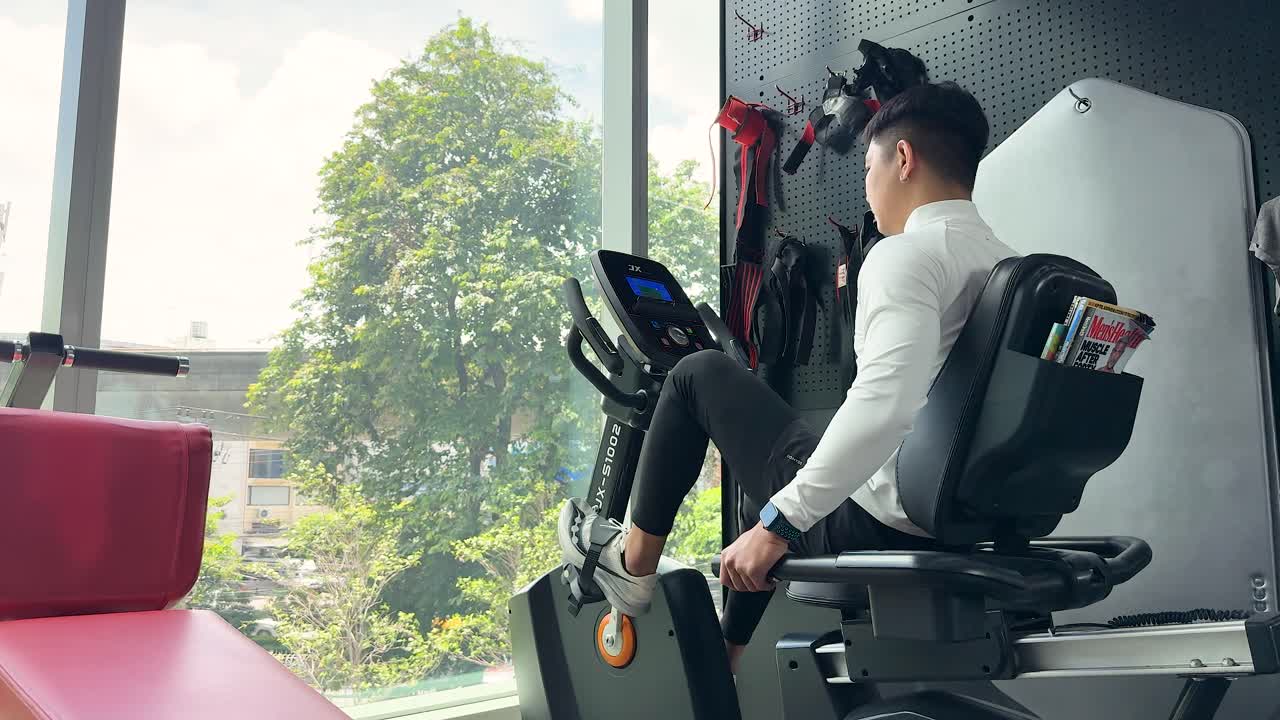 A man uses a recumbent bike in a well-lit gym with large windows and a view of trees