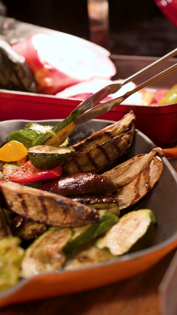 Pinze metalliche selezionano zucchine e peperoni arrostiti da un vassoio da buffet colorato sotto un'illuminazione calda.