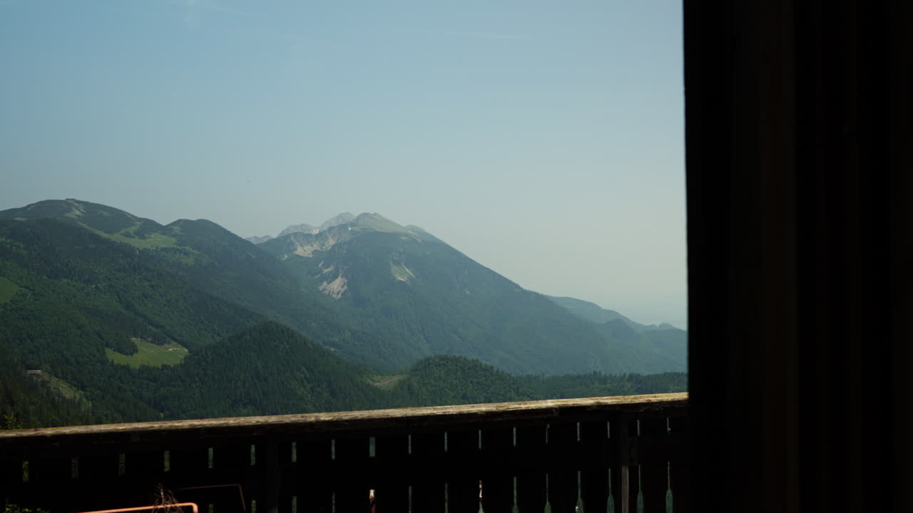 Cinematic view looking out of a window at Koča na Golici hut in the Karavanke mountains, Slovenia. The shot reveals peaceful alpine scenery, mountain slopes, and lush green landscape.