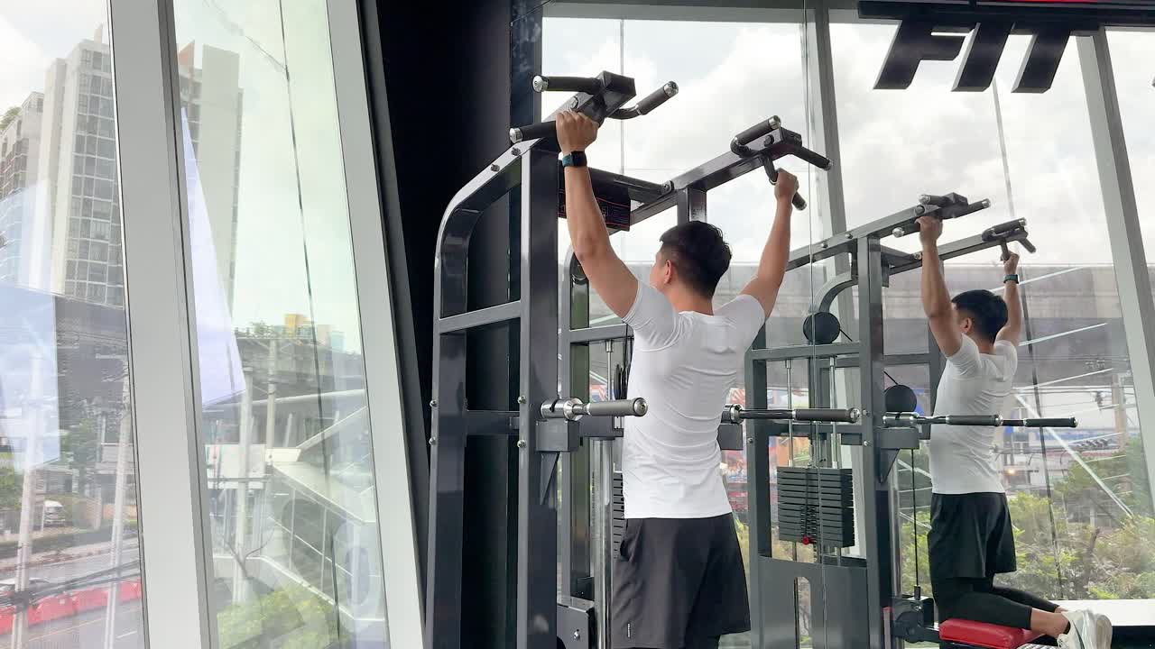 Man exercises on pull-up machine in a bright, modern gym with large windows and city views