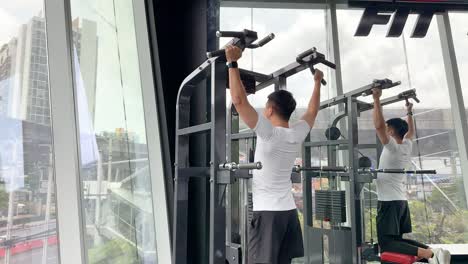 man exercises on pull-up machine in a bright, modern gym with large windows and city views