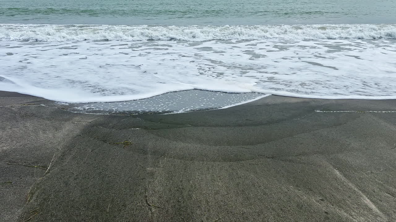 Onde si infrangono su una spiaggia di sabbia grigia sotto un cielo nuvoloso durante le ore diurne