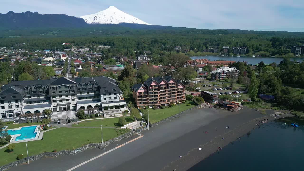 Orbit drone shot of lakeside hotels, black sand beach, and the snow-capped Villarrica Volcano in the background, captured in Pucón, Chile