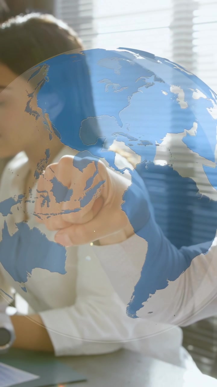 Video vertical: Mujer manejando documentos en el escritorio mientras el brazo de un colega entra, tocando un globo terráqueo que muestra datos empresariales.