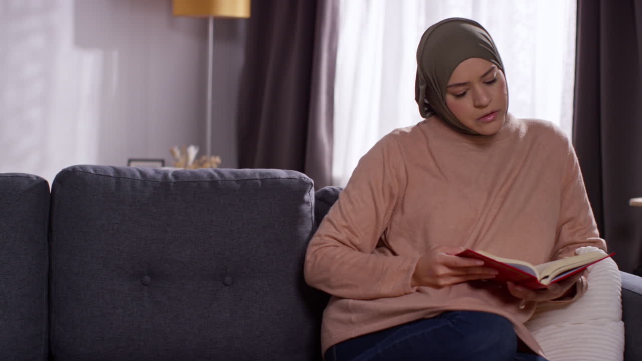 Donna musulmana con l'hijab seduta sul divano a casa che legge o studia il Corano 1