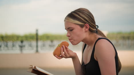 mujer con camiseta negra sentada al aire libre, disfrutando de una hamburguesa mientras lee un libro, con una valla decorativa y un cielo claro y brillante en el fondo, participando en la lectura casual y comiendo al aire libre