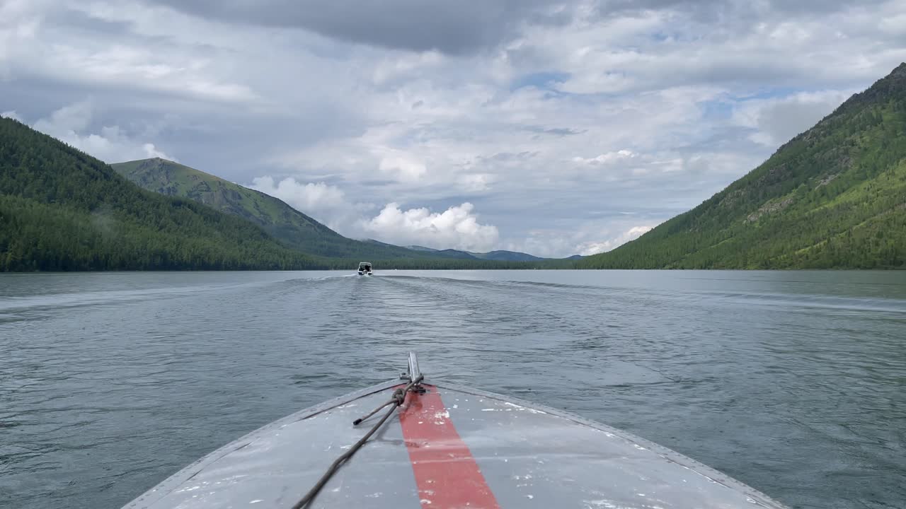 POV von einem fahrenden Boot, das die Multinsky-Seen in der Republik Altai, Russland, überquert, mit bewaldeten Bergen auf beiden Seiten und einem dramatischen bewölkten Himmel über ruhigem Wasser.