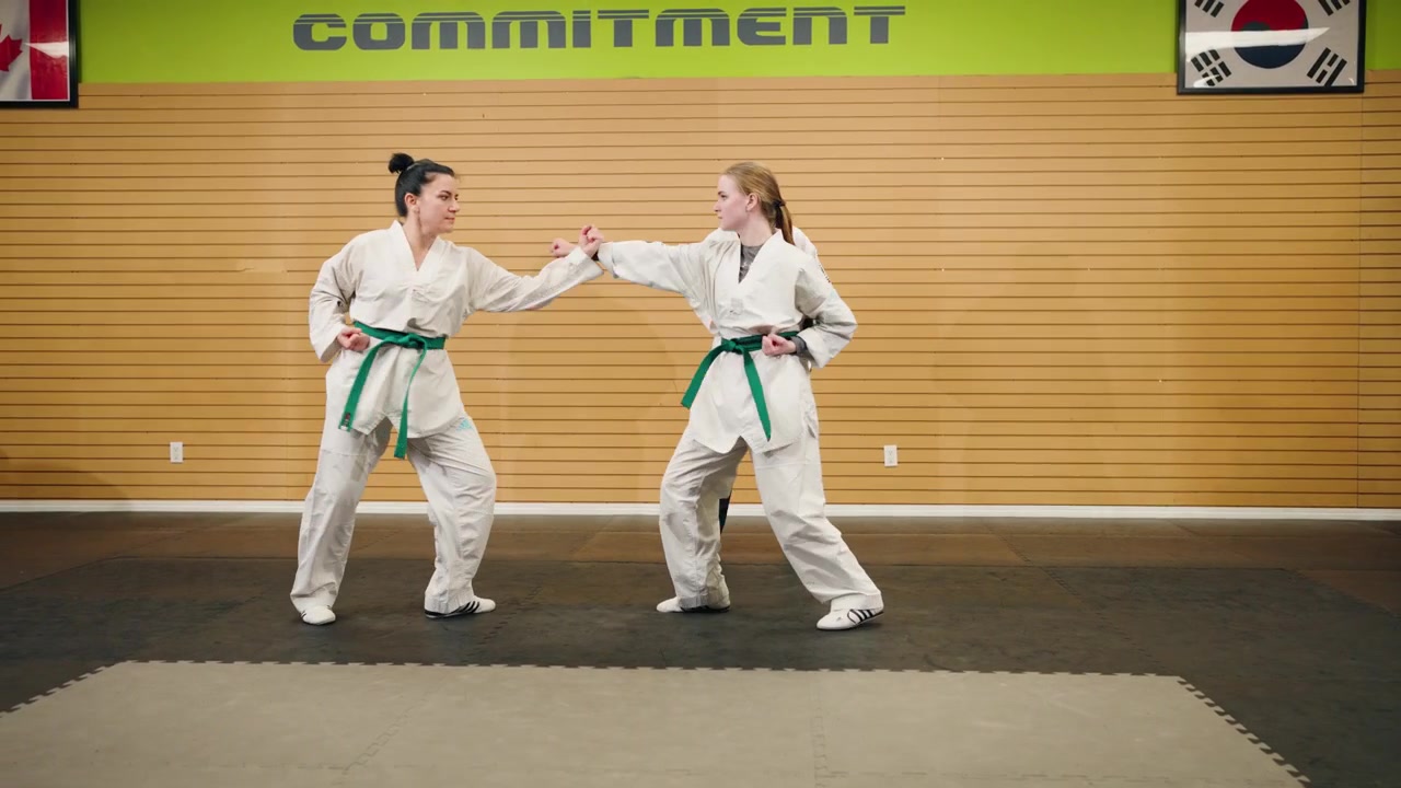 Góc rộng: Hai nữ thiếu niên thực hiện chuỗi động tác kết hợp tập luyện Taekwondo, Karate, Kung Fu (đá, đấm, vật ngã) cùng võ sư đai đen bên trong một trung tâm thể hình.