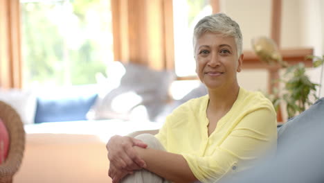 Portrait-of-happy-senior-biracial-woman-sitting-on-couch-at-window-at-home,-slow-motion