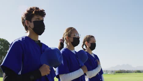 Diverse-team-of-female-baseball-players-in-face-masks-standing-in-line-with-hands-on-hearts