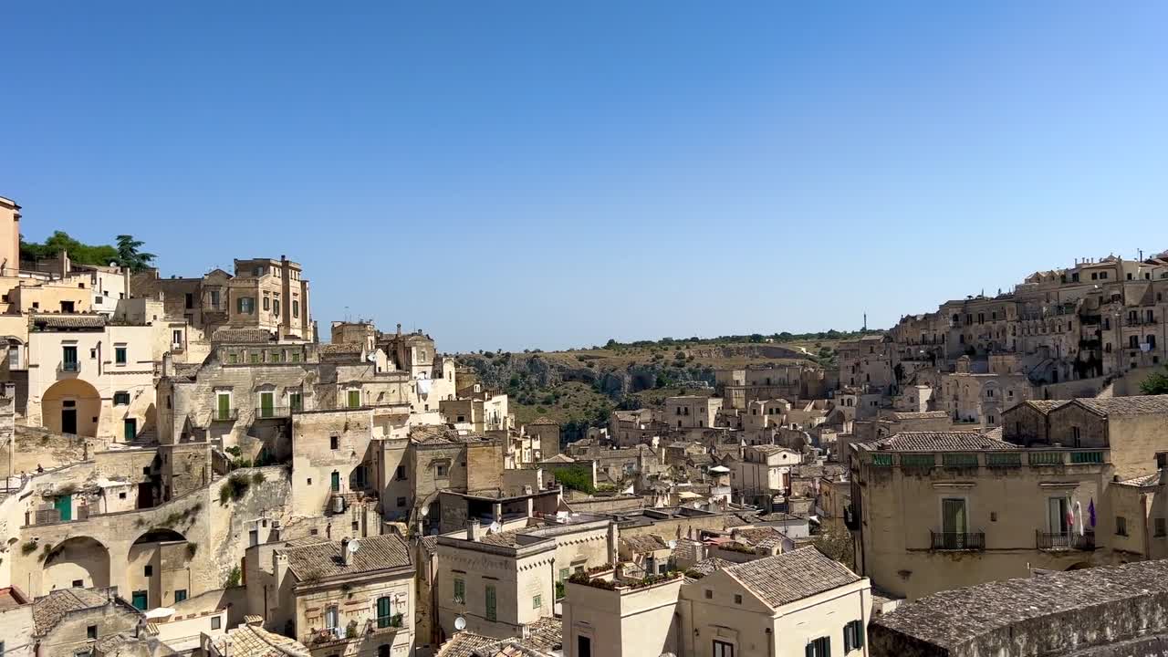 Vista aerea con drone di una città storica collinare caratterizzata da edifici in pietra raggruppati strettamente con strutture simbolo color pesca sotto un cielo azzurro nel sud Italia