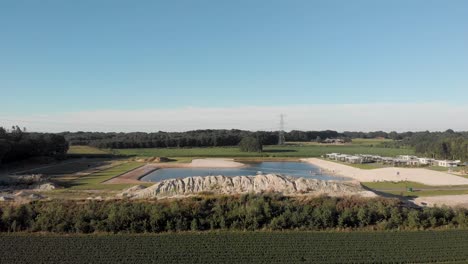 aerial reveal of recreational pond and summer homes in farmland surrounding with a field of recently headed lilies and a large sand hill in the foreground against a clear blue sky