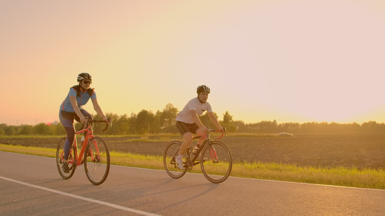 Handsome bearded professional male cyclist riding his racing bicycle in the morning together with his girlfriend both wearing protective helmets and eyeglasses sun shining through between them