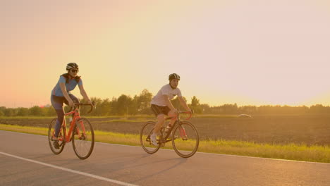 Handsome-bearded-professional-male-cyclist-riding-his-racing-bicycle-in-the-morning-together-with-his-girlfriend-both-wearing-protective-helmets-and-eyeglasses-sun-shining-through-between-them