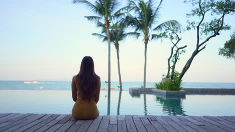 a woman with her back to the camera sits on a wooden jetty while looking out on the resort infinity pool and ocean horizon