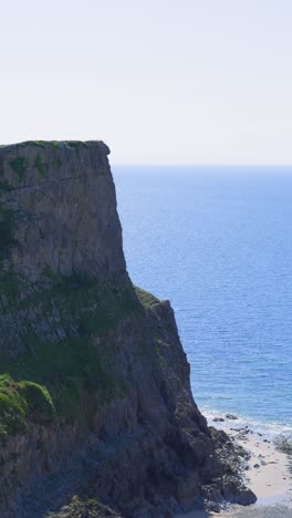 vertical video of steep grass covered cliff top along sea coastline with sandy beach down below and calm rolling waves in gower, wales uk.