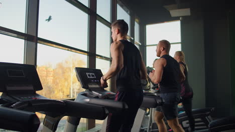 Dos-Hombres-Corriendo-En-Una-Cinta-De-Correr-Al-Fondo-De-Una-Gran-Ventana-En-El-Gimnasio.-Hombres-Jóvenes-Corriendo-En-El-Gimnasio.-Entrenamientos-Aeróbicos-En-El-Gimnasio.