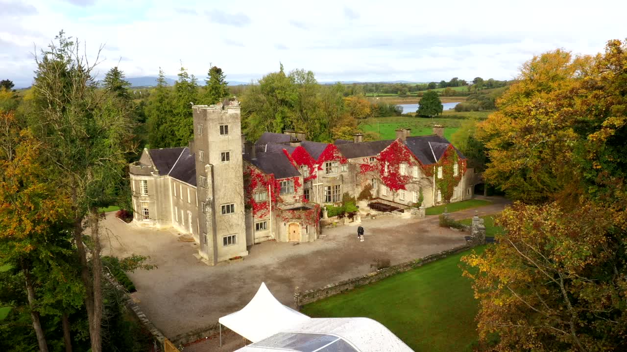 Sunset drone approach through trees of Belle Isle Castle showing golden hues over towers and estate grounds
