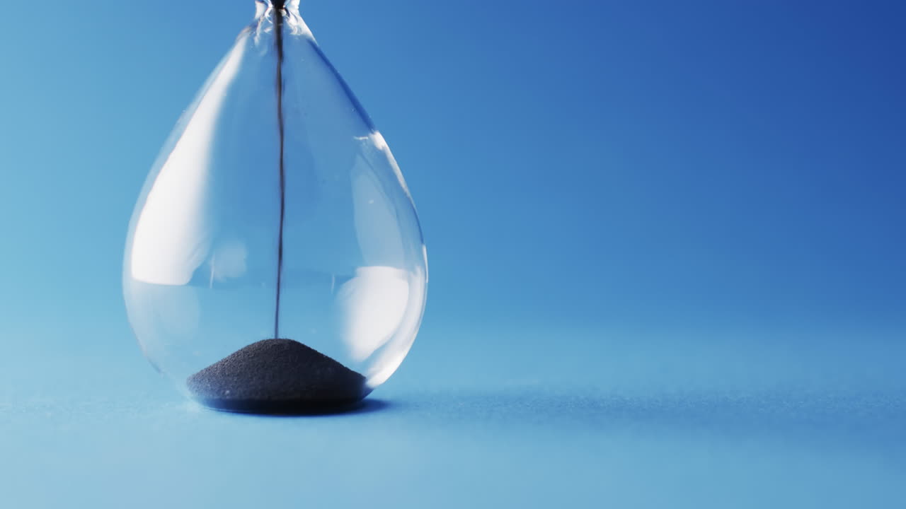Video of close up of hourglass with black sand pouring with copy space on blue background