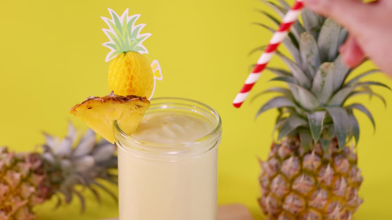 Hand stirs creamy pineapple smoothie in glass with garnish, bright yellow background, natural lighting