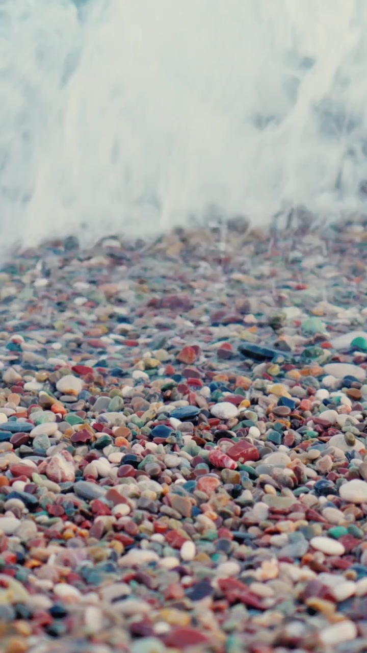 Gentle ocean waves roll onto a pebble beach with foamy water and natural tones, filmed in high quality vertical video for mobile and social media.