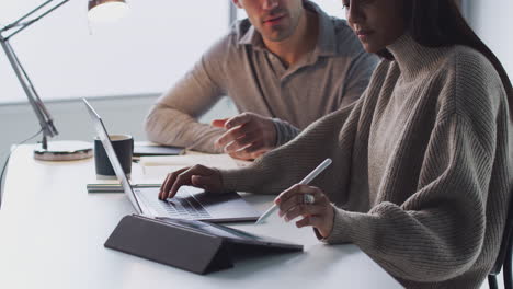 businesswoman working on laptop at desk with male colleague using digital tablet and stylus