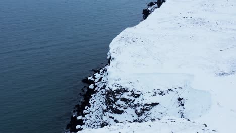 Acantilados-Cubiertos-De-Nieve-Blanca-A-Lo-Largo-De-Aguas-Tranquilas-Del-Océano,-Islandia,-Antena