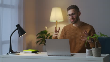 Un-Joven-Vestido-Con-Un-Suéter-Marrón-Da-La-Bienvenida-A-Los-Interlocutores-En-Una-Conferencia-En-Línea-De-Video-Chat-De-Colegas-Que-Trabajan-Desde-Casa