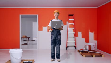 cuerpo lleno de pintor hombre asiático con casco de seguridad usando una computadora portátil y sonriendo mientras está de pie con la pared de pintura en la habitación