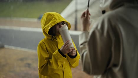 Por-Encima-Del-Hombro,-Una-Niña-Feliz-Con-Una-Chaqueta-Amarilla-Se-Comunica-Con-Su-Madre-Y-Bebe-Agua-De-Una-Botella-Blanca-En-El-Parque-Durante-Una-Caminata-Y-Una-Fuerte-Lluvia.