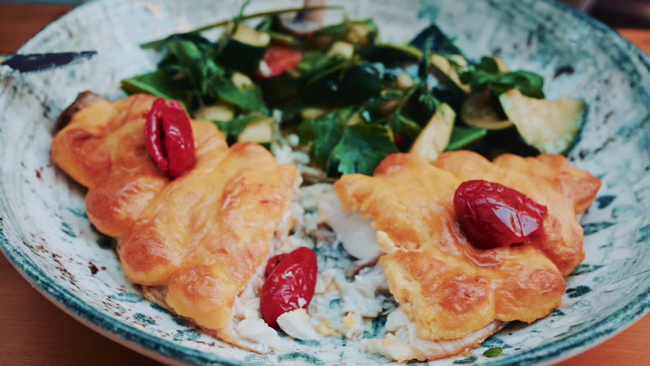 Close up of the flaky pastry on top of a fish filet, topped with red peppers, with a fork cutting into it, vegetables in the background