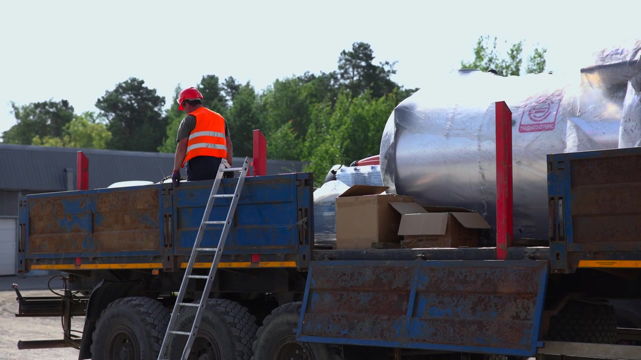 Worker in red hard hat and orange safety vest bends over flatbed truck beside aluminum ladder, reaching toward cargo in practical industrial yard with wrapped machinery, cardboard boxes, blue metal
