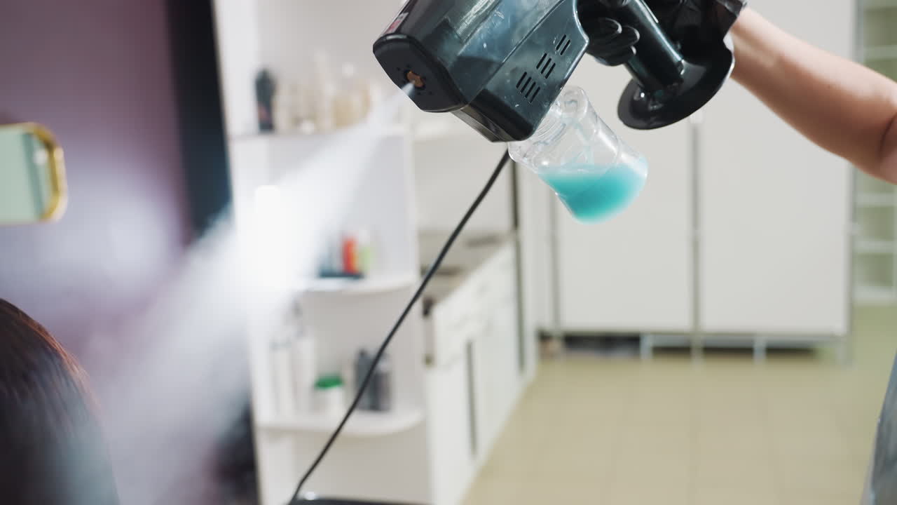 미세한 증기를 분사하는 휴대용 분무기가 미용실 의자 전체에 분사되고, 파란색 용액 병이 부착되어 있으며, 장갑을 낀 기술자가 방아쇠를 작동시키고, 현대적인 미용실 인테리어와 부드러운 트리트먼트 분위기가 연출됩니다.