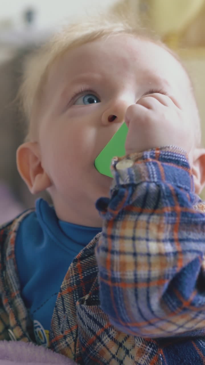 funny blue eyed baby in checkered shirt nibbles green cube leaning on soft bed in light children room close view