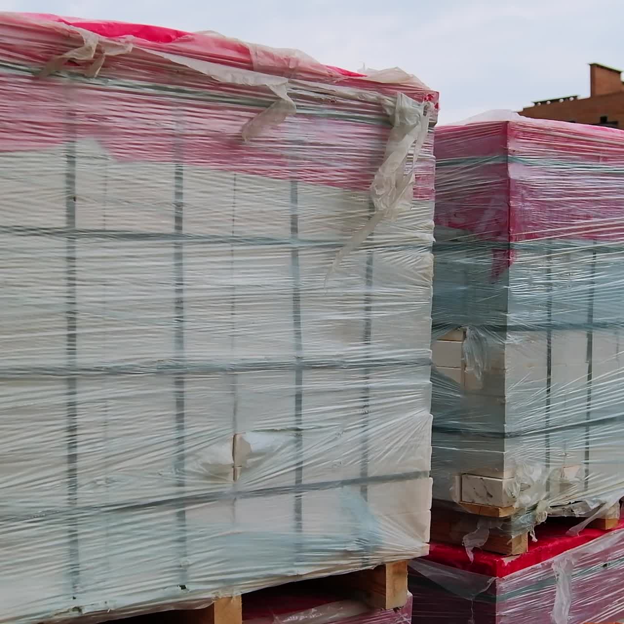 Big piles of white bricks in stacks covered with plastic. Multistoried building in the construction process at backdrop
