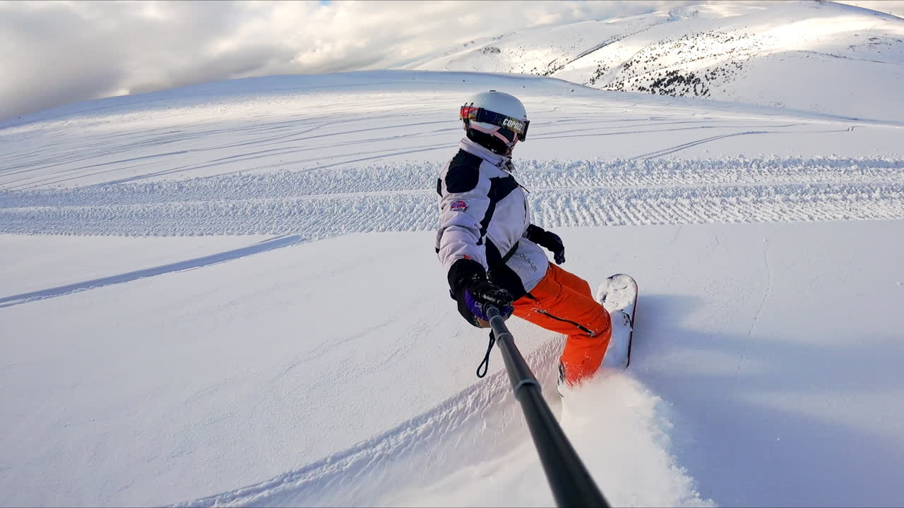 Sportsman wearing a white-and-black jacket and orange pants takes selfie video. Snowboarder stops at the grader's trail.
