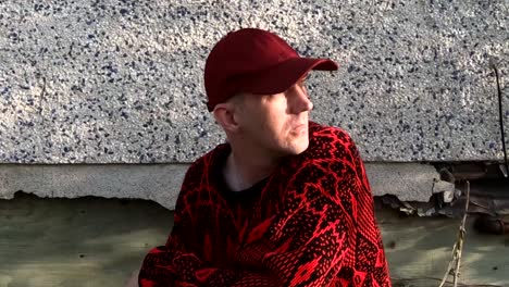 adult male wearing a black and red sweater and hat in the wind against the side of a house