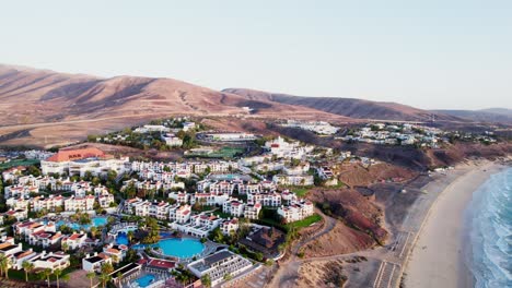 coastal resort in fuerteventura with white buildings and pools, flanked by hills and a beach