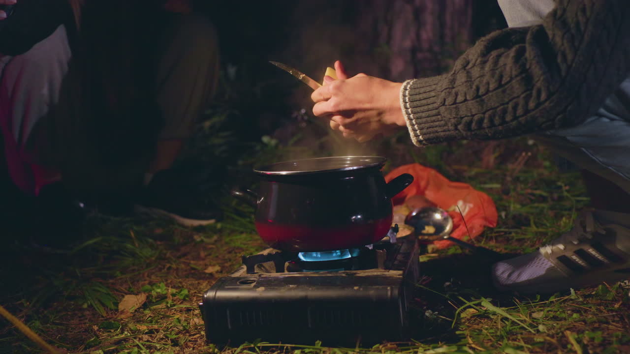 Close up of people cooking during night camping in forest as one person peels food over steaming pot on portable stove while other assists with light, surrounded by grass