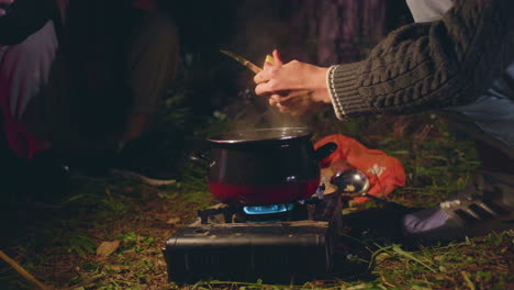 close up of people cooking during night camping in forest as one person peels food over steaming pot on portable stove while other assists with light, surrounded by grass
