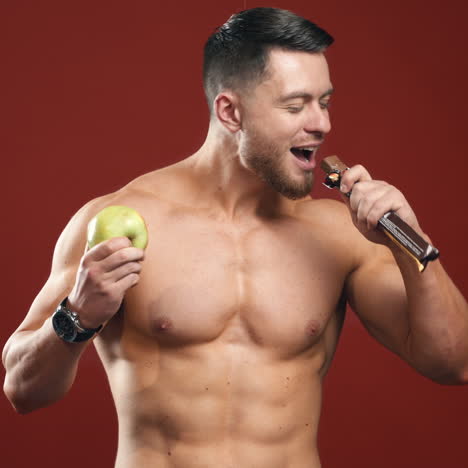 sportsman choosing vitamins. muscular athlete holding sweet chocolate bar and healthy apple. shirtless bodybuilder eats fresh apple on red studio background.
