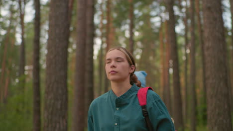 dama con mochila roja y camisa verde camina a través de un sendero del bosque sereno, rodeado de árboles altos y naturaleza pacífica, su amiga, con una bufanda azul, camina detrás de ella, mirando hacia abajo en pensamiento