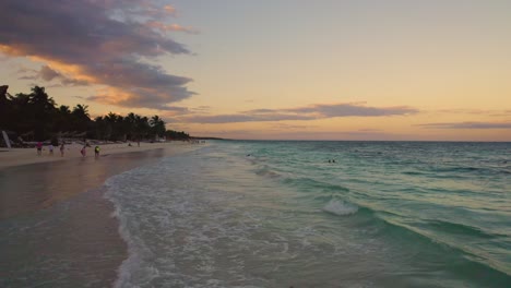 Vuelo-Bajo-Aéreo-Sobre-Las-Olas-Del-Mar-Caribe-En-La-Playa-De-Akiin-En-Tulum,-México-Durante-La-Puesta-De-Sol-Con-Gente-Disfrutando-De-Sus-Vacaciones-En-La-Playa